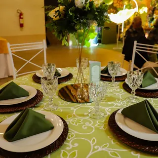 Mesa dos convidados com toalha verde estampada, pratos brancos, guardanapos verdes e arranjo de rosas