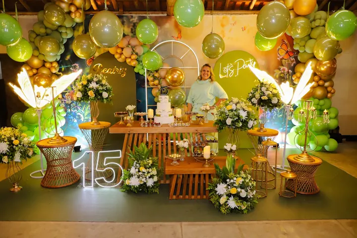 Patrícia sorrindo ao lado da mesa do bolo de uma festa de quinze anos decorada em verde e dourado, com balões, flores e letreiro com o nome Beatriz