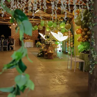 Salão da festa visto da entrada, com cortina de flores brancas e balões verdes e dourados ao fundo