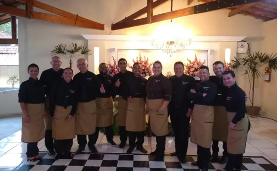 Equipe da Patrícia Festas, 14 pessoas de uniforme preto e avental bege, num salão de festas decorado com flores